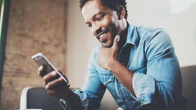 Happy bearded African businessman using phone while sitting on sofa at his modern home.Concept of young people working mobile devices.Blurred background